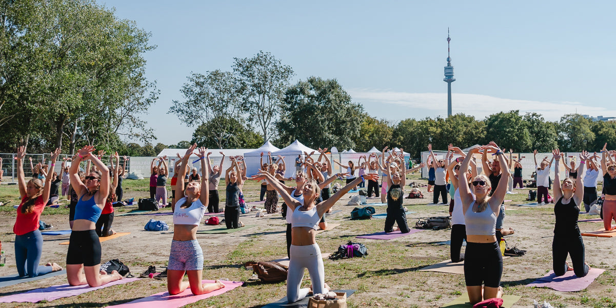 A group of people practicing yoga outdoors in a field during the Yoga Junkies Festival. They are in various poses, surrounded by nature and the festival atmosphere.